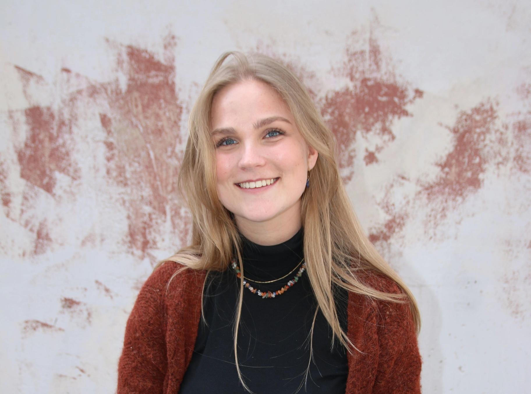 A smiling young woman with blonde hair wears a black turtleneck and a rust-colored cardigan against a textured wall.