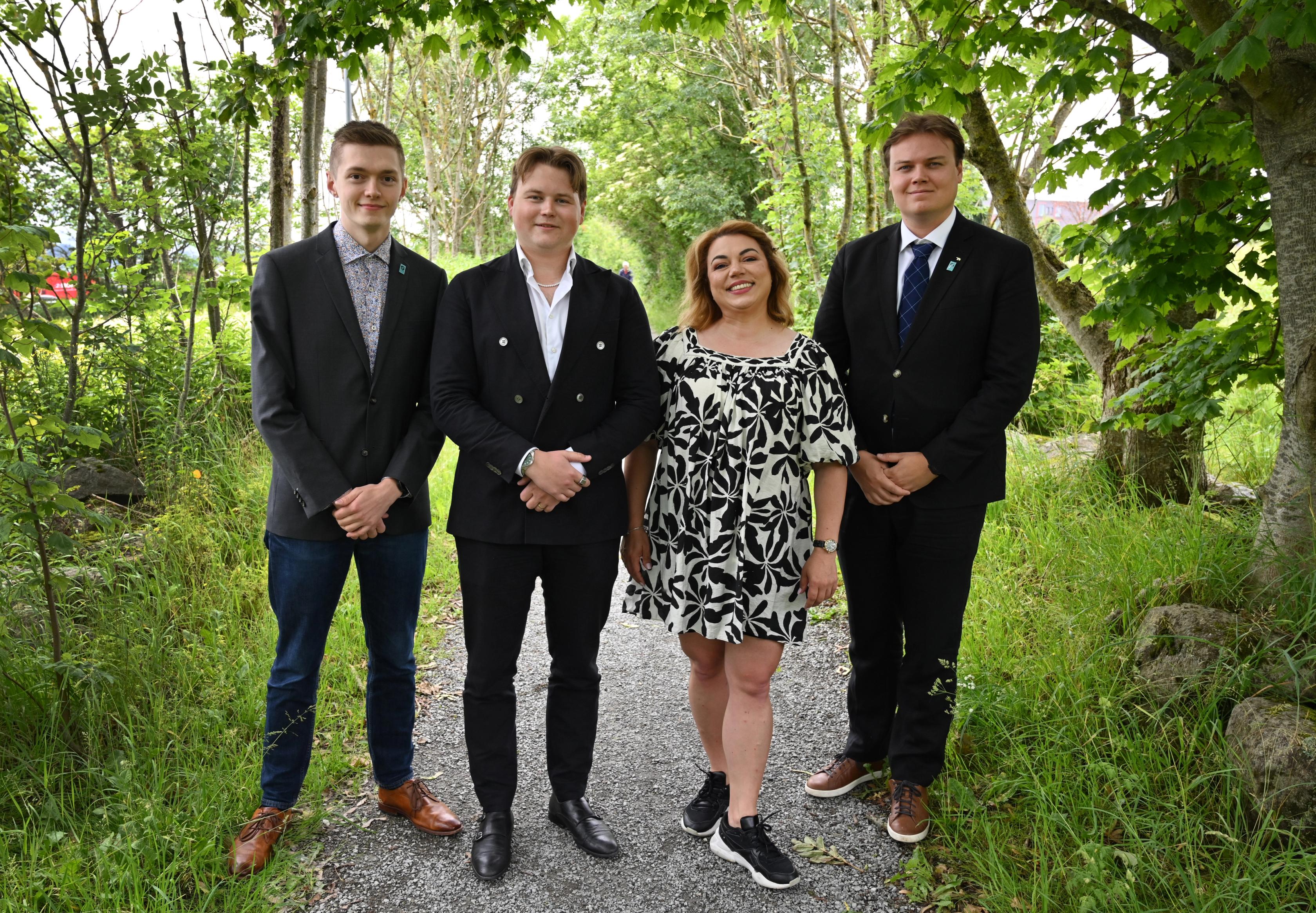 Four people, three men and one woman, stand on an outdoor path flanked by trees.
