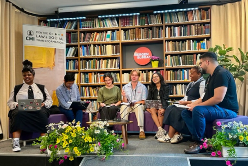A diverse panel of seven people sits on a stage in front of a bookshelf, with "Centre on Law & Social Transformation" and "Bergen Global" banners visible.