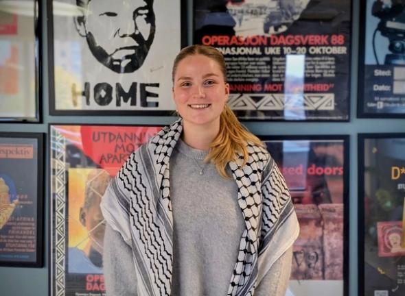 A young woman smiling, wearing a grey sweater and a black and white keffiyeh, standing in front of a wall covered with framed posters.