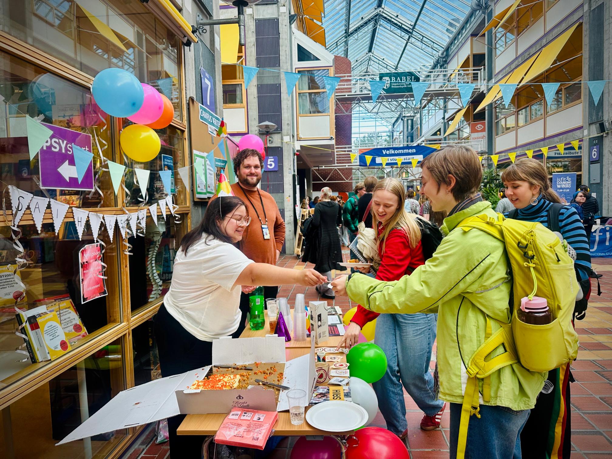 Young adults at an event booth in a bright atrium, a woman hands out items from a table with pizza and balloons.