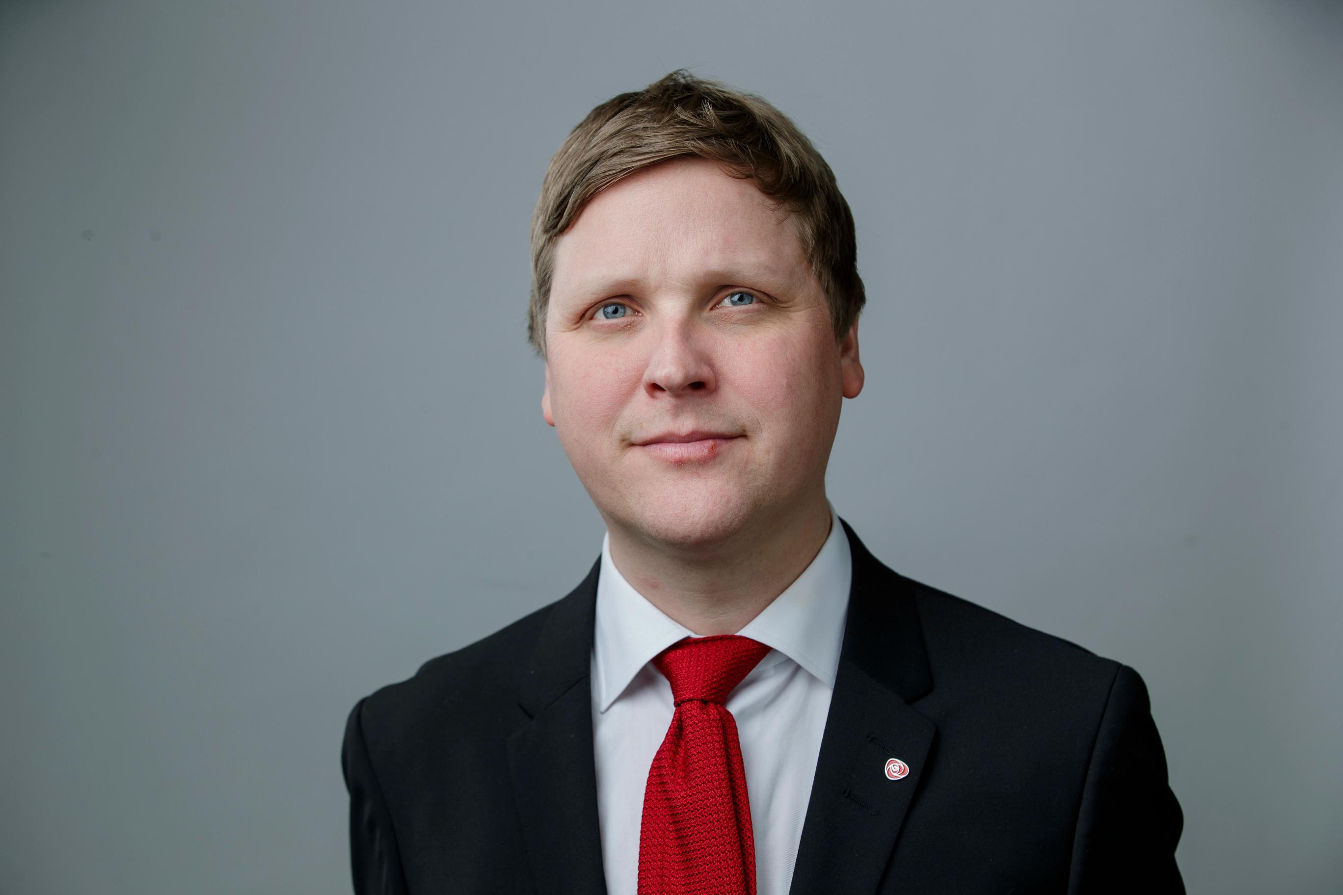 A man with light hair and blue eyes, wearing a black suit and red tie, smiles subtly against a grey background.