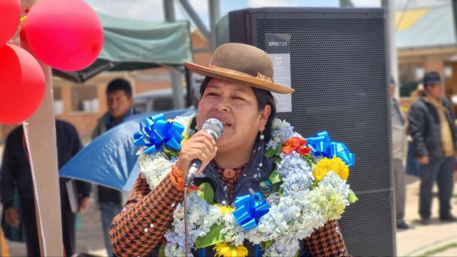 A woman wearing a hat and flower garland holds a microphone in her hand.