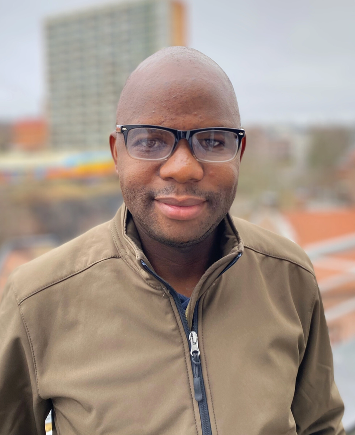 a man wearing glasses and a brown jacket smiles for the camera