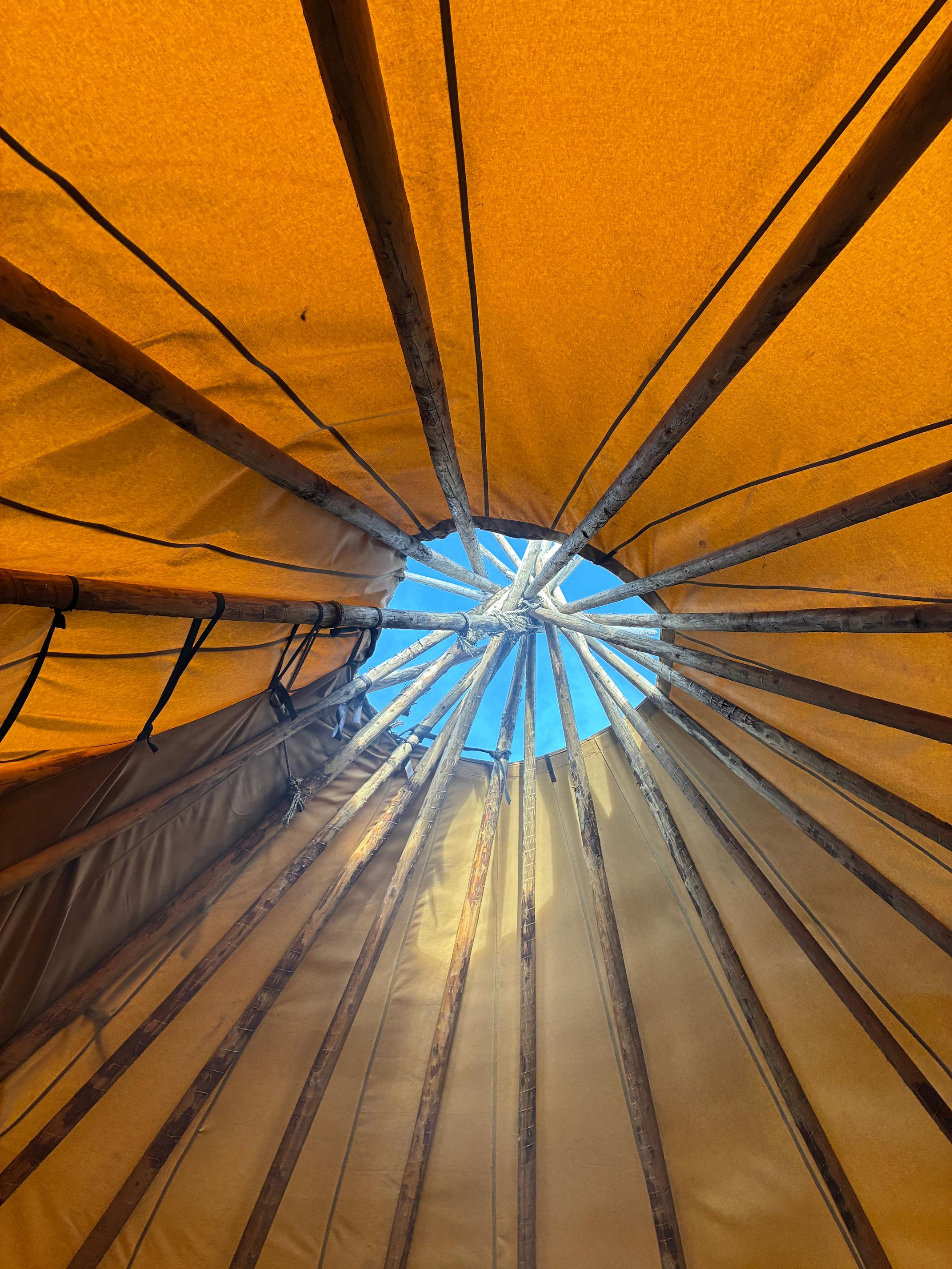 Inside a teepee, orange fabric walls and converging wooden poles frame a blue sky through the top opening.