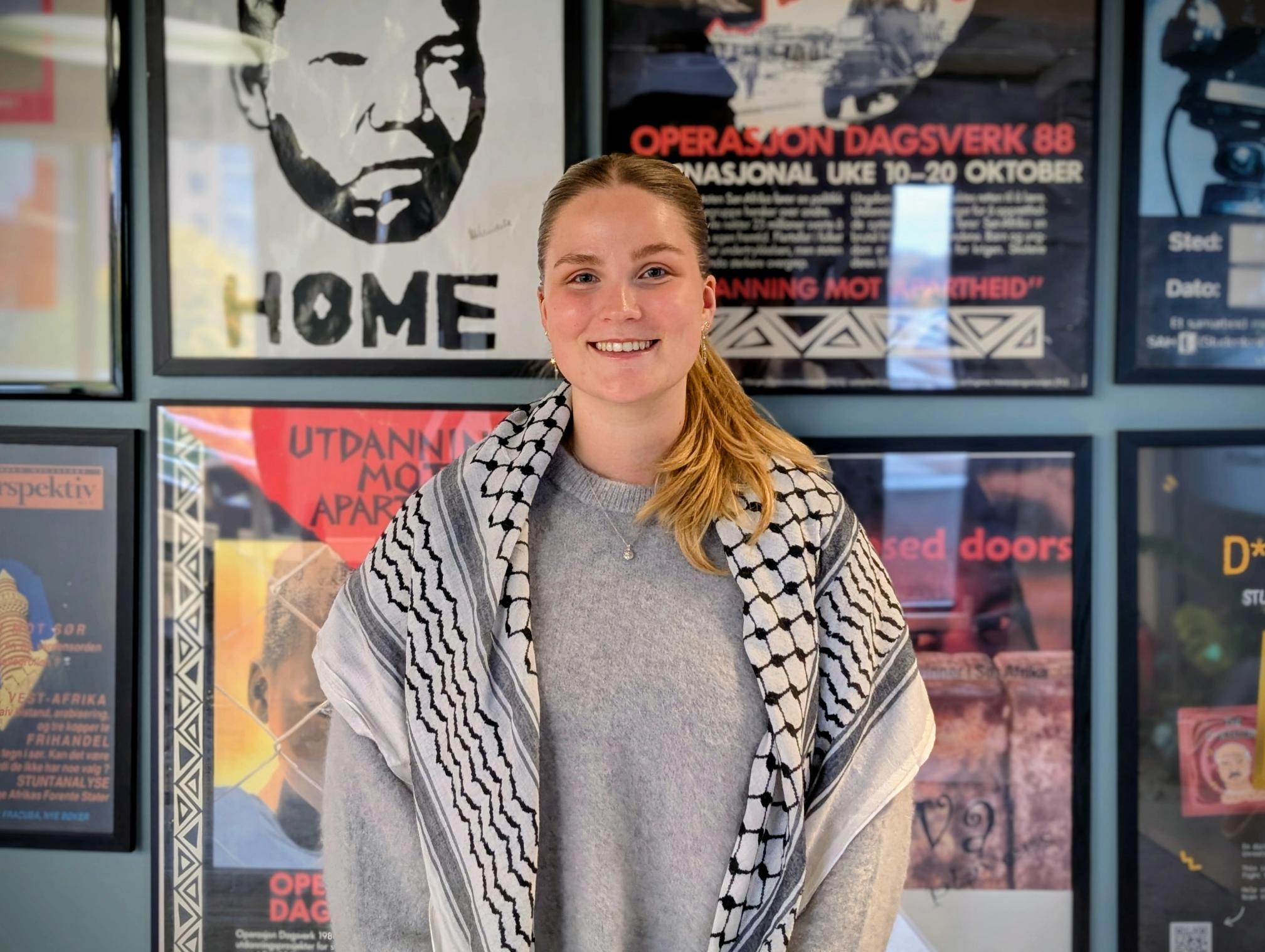 A smiling young woman wearing a black and white keffiyeh stands in front of posters, one depicting Nelson Mandela.