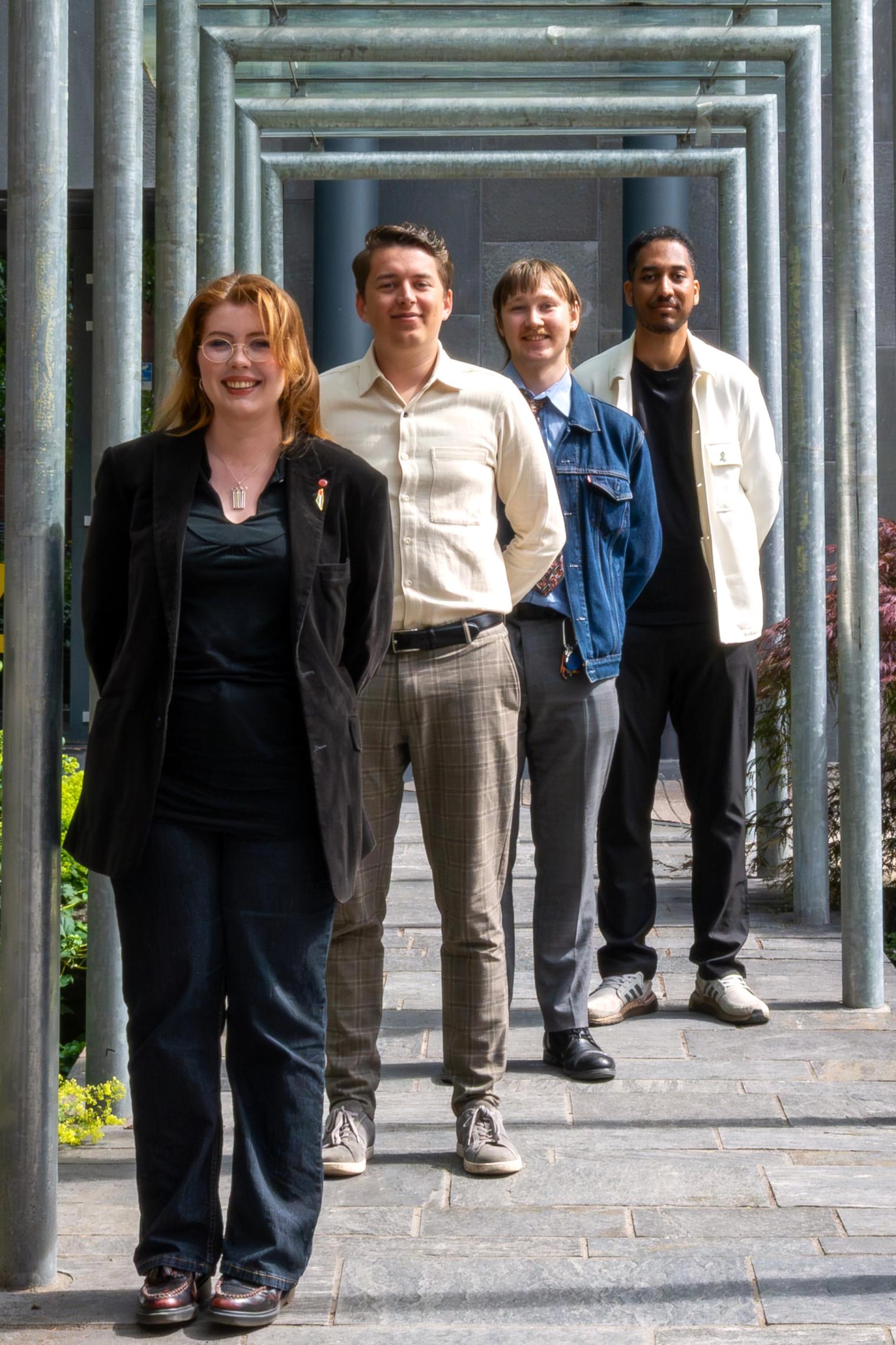 Four smiling young adults stand in a line under a metal archway.
