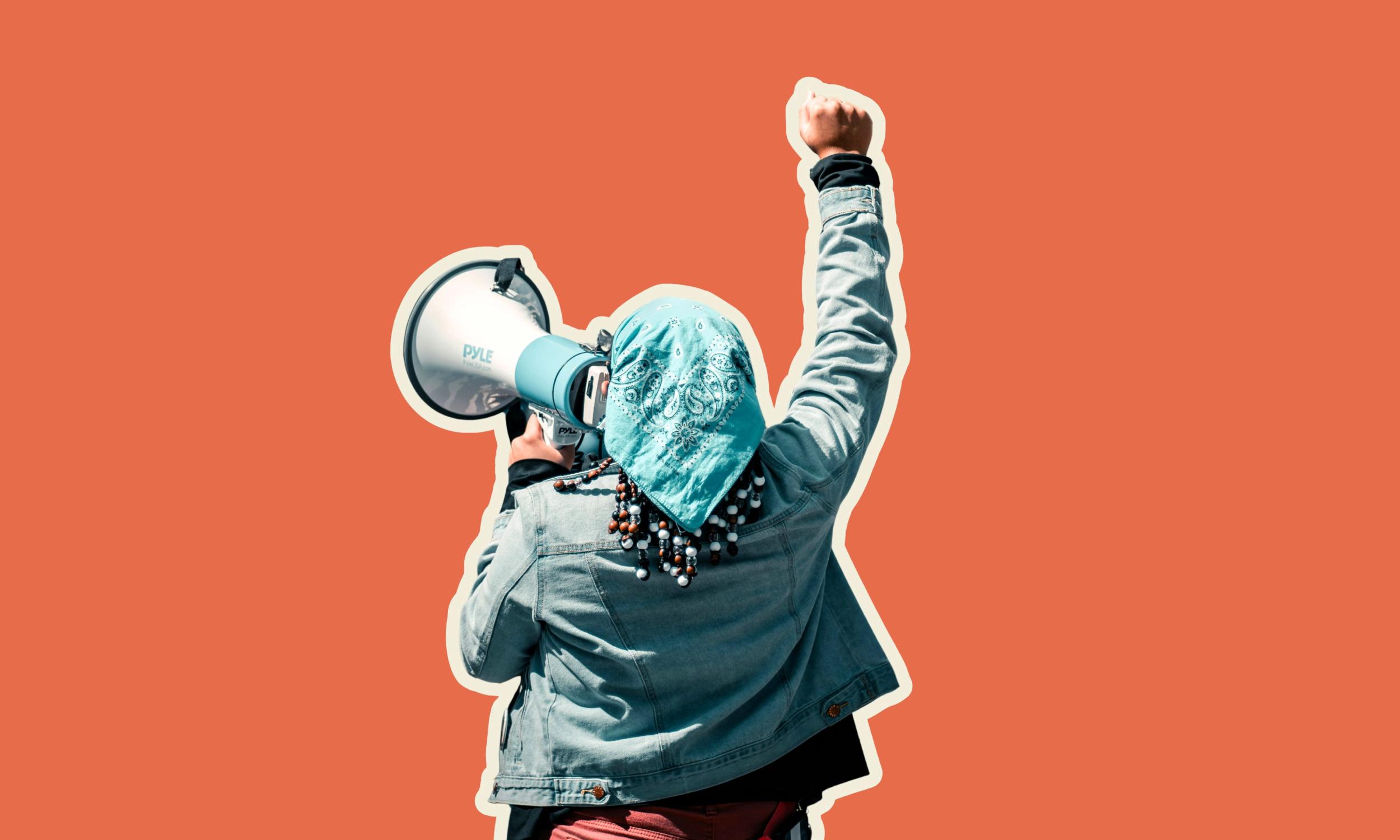 Person from behind, wearing a bandana and denim jacket, holding a megaphone and raising a fist.
