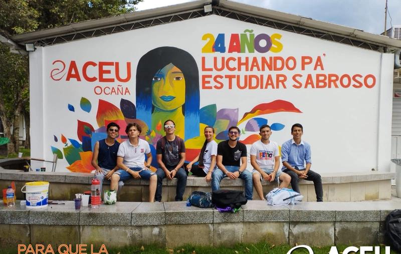 Students from ACEU's local chapter in Ocaña sit outside a building with a freshly painted ACEU logo on it.