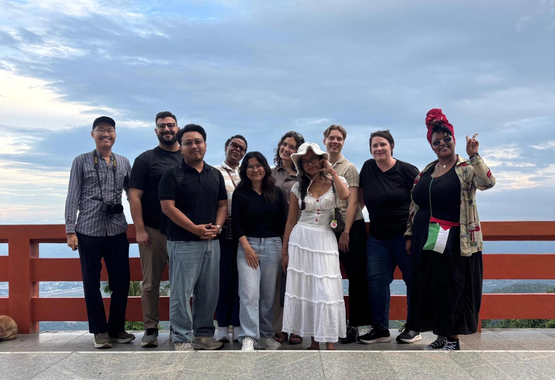 A diverse group of ten people smiling and posing outdoors on a viewing platform with a cloudy sky and distant landscape.