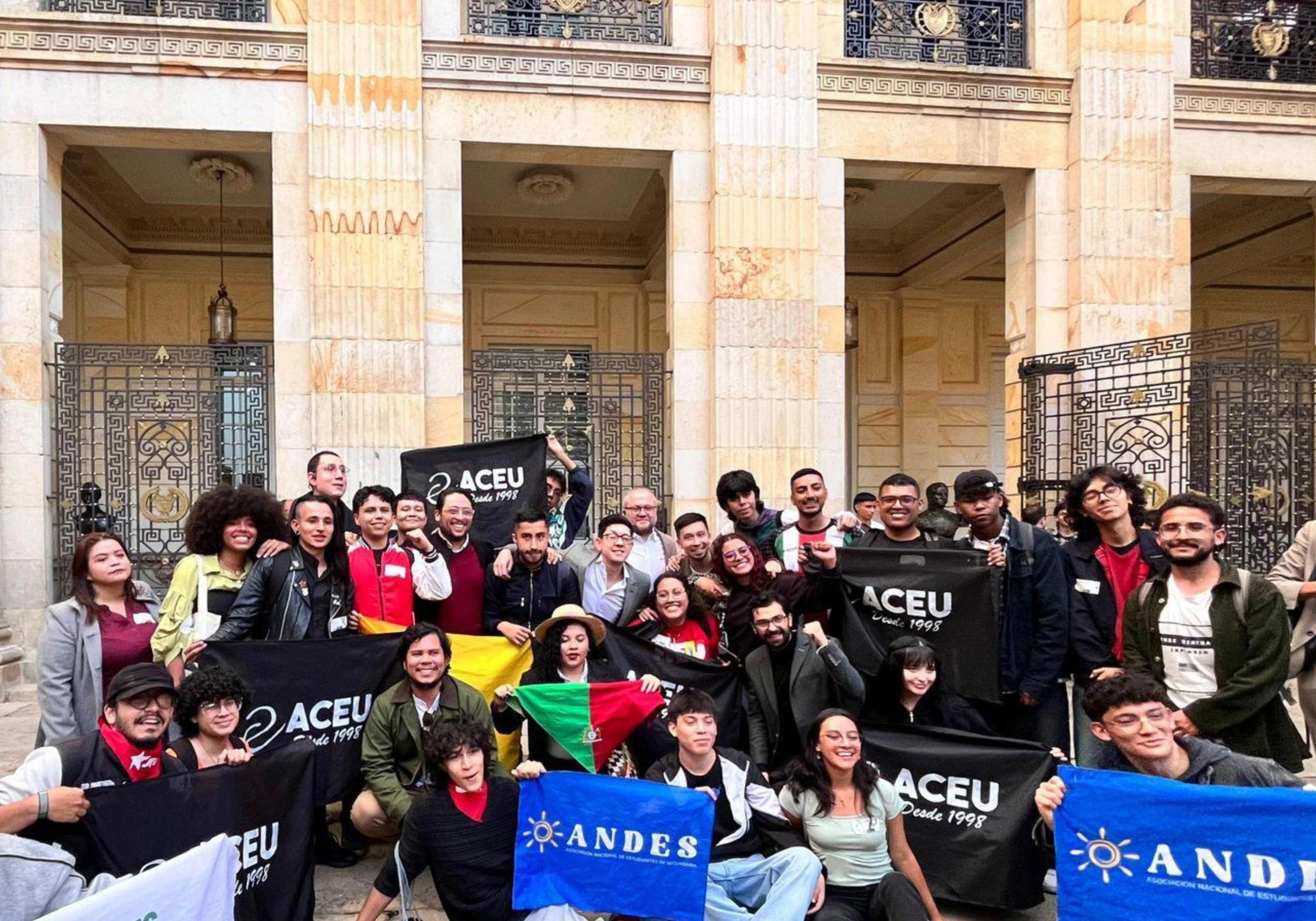A diverse group of smiling people hold banners for ACEU and ANDES, along with a Palestinian flag, in front of an ornate stone building.