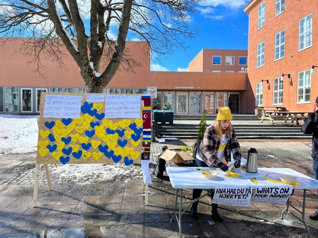 En kvinnelig student som står på stand med et skilt "hva har du på hjertet?" I bakgrunnen en tavle med hjerter i gult og blått hvor studenter har delt sine tanker om krigen i Ukraina. 