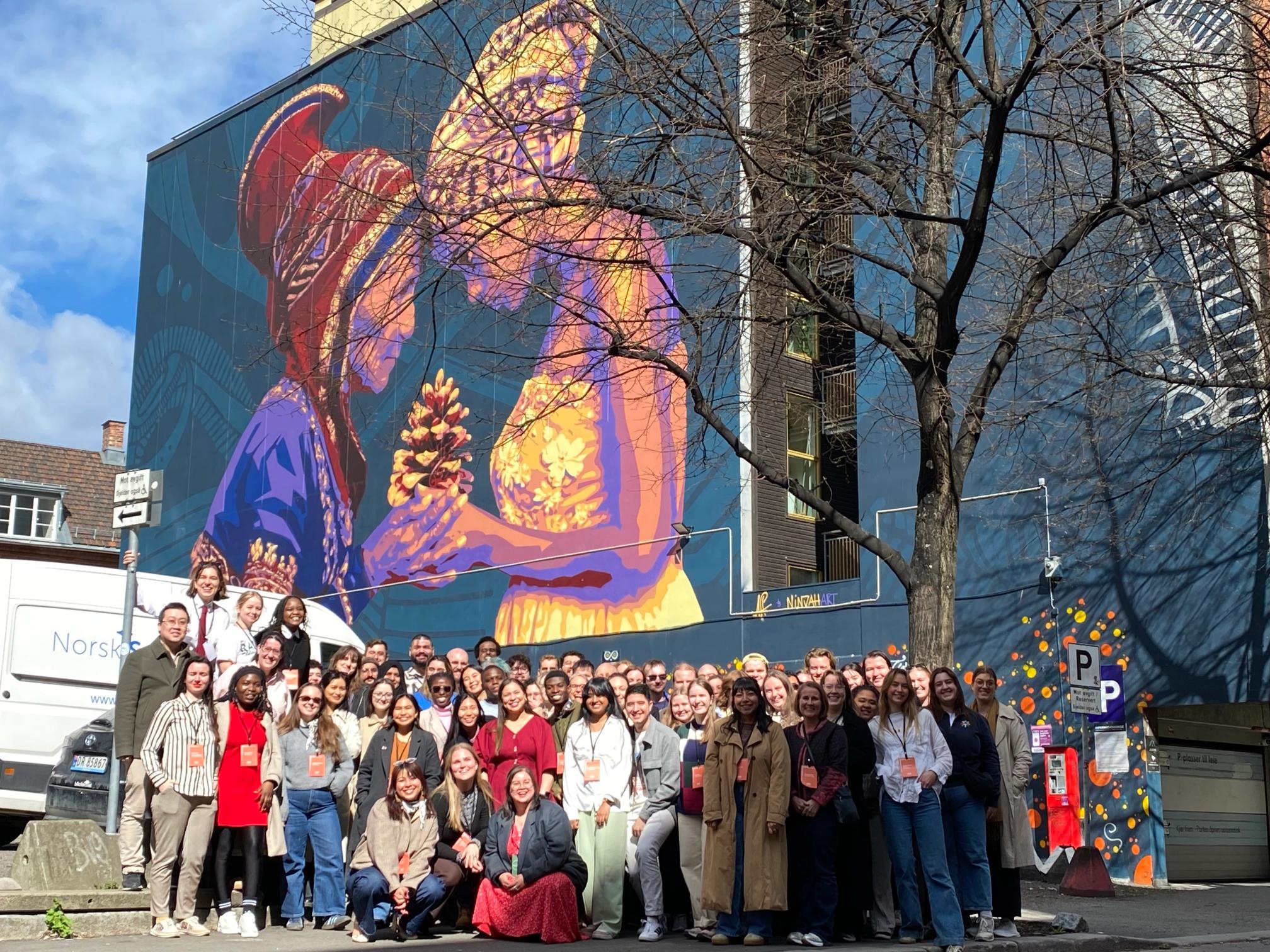 A large group of people stands in front of a vibrant mural depicting two stylized figures, one in purple and one in gold, holding a pinecone.