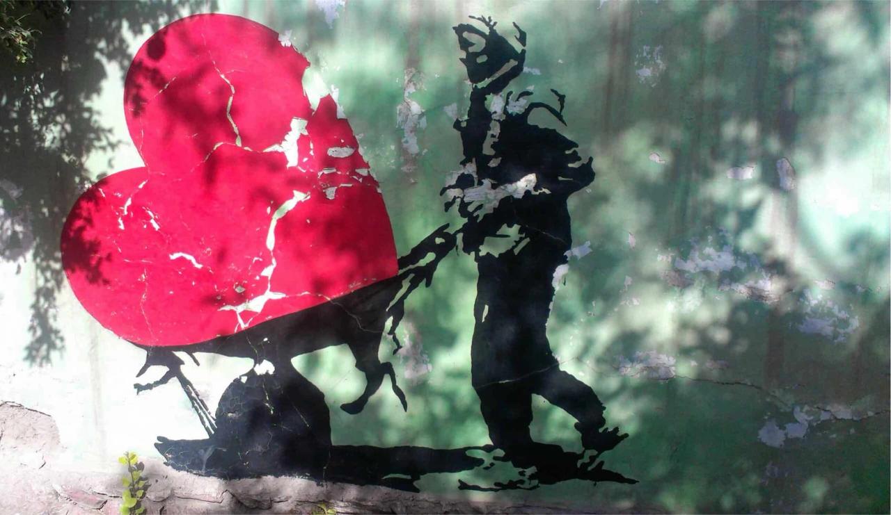 Stencil art of a dark silhouette pulling a cracked red heart on a distressed green wall, dappled with tree shadows.