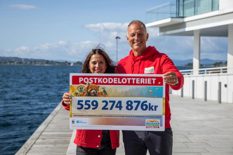 Two smiling people hold a large Postkodlotteriet check displaying 559,274,876 kr against a sunny waterfront.