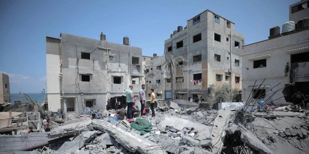 A group of people stands on rubble in front of damaged buildings overlooking the sea.