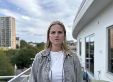 A blonde woman wearing a grey denim jacket stands on a balcony with a city view.