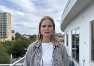 A young woman in a grey jacket stands on a balcony with a city skyline behind her.
