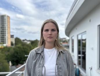 A young woman with blonde hair in a grey jacket stands on a balcony overlooking a city.