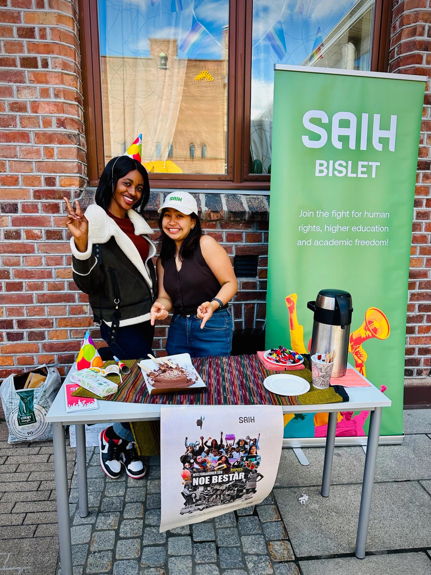 Two smiling women, one in a party hat, stand behind a table with a chocolate cake and a green SAIH Bislet banner promoting human rights.