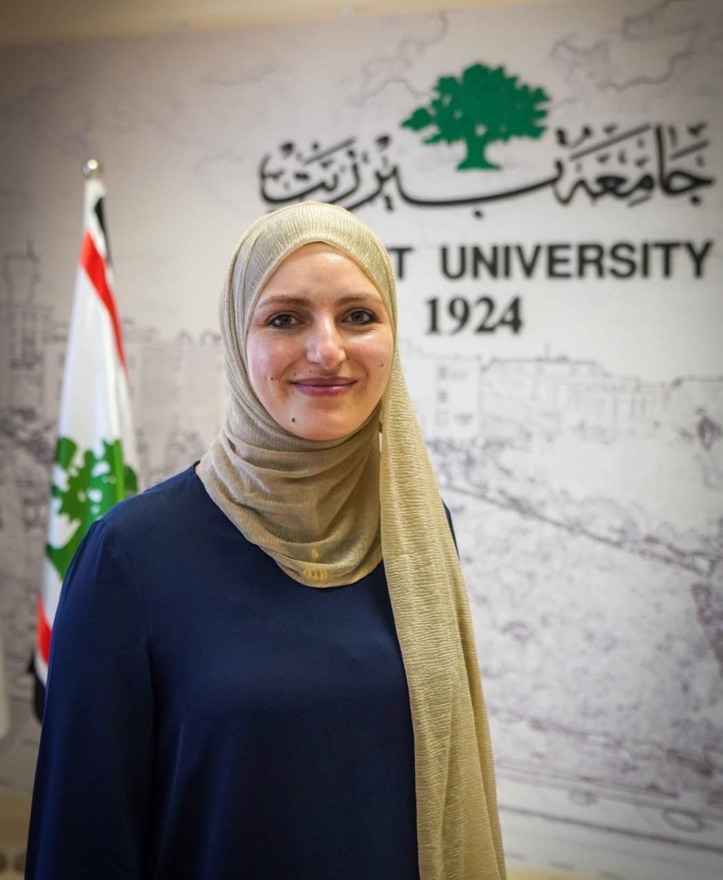 Smiling woman in a gold hijab and navy top stands before a Beirut University logo and a flag.