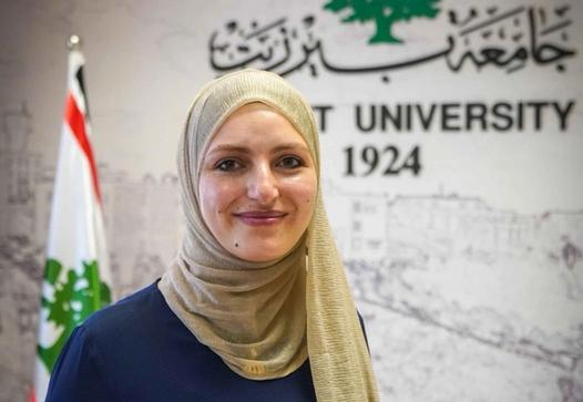 Smiling woman in a gold hijab and navy top stands before a Beirut University logo and a flag.