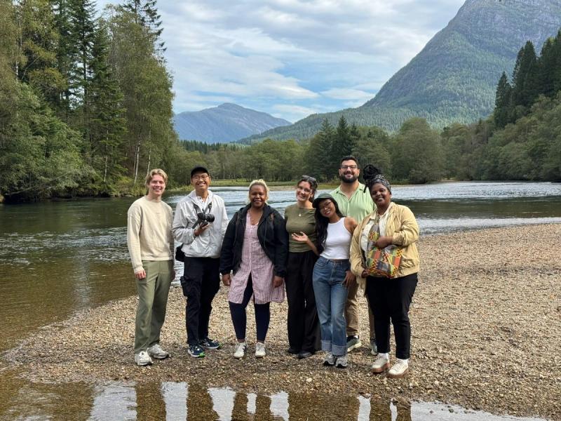 A diverse group of eight people stand on a rocky riverbank with mountains and trees in the background.