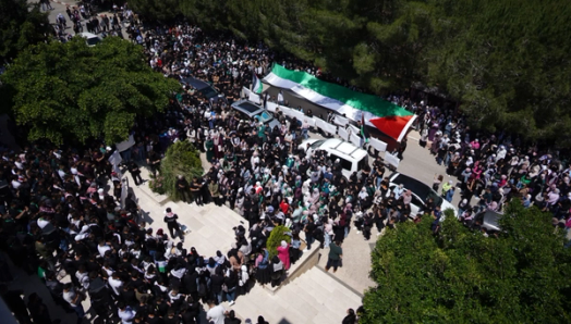 An aerial view of a large procession with a huge Palestinian flag draped over a white object, surrounded by a dense crowd of people, cars, and trees.