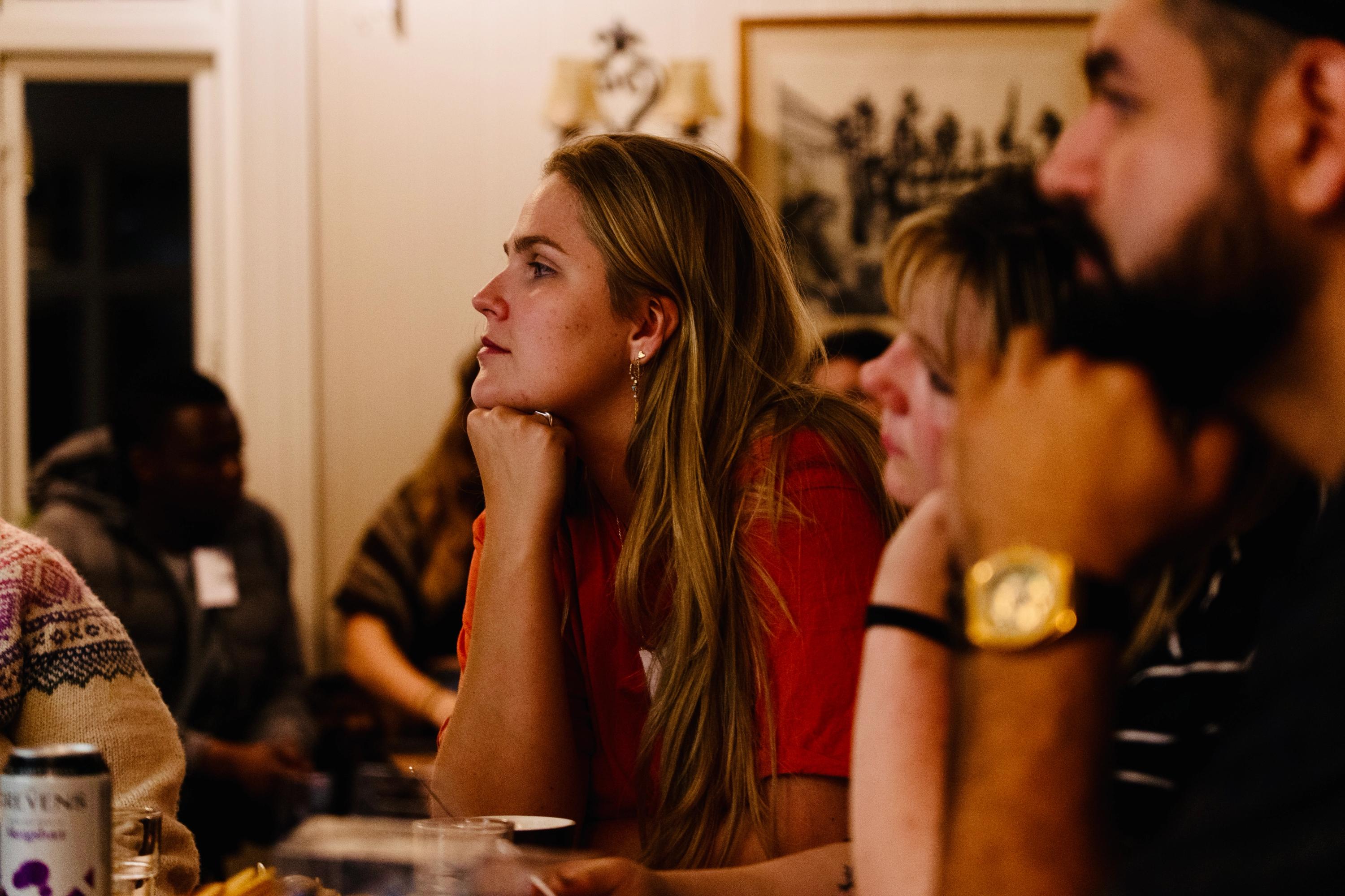 A blonde woman in a red shirt listens intently, chin resting on her hand, surrounded by other people.