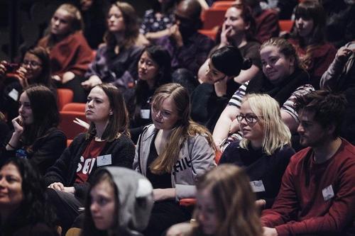 Et publikum lytter oppmerksomt til en debatt på scenen foran seg. 