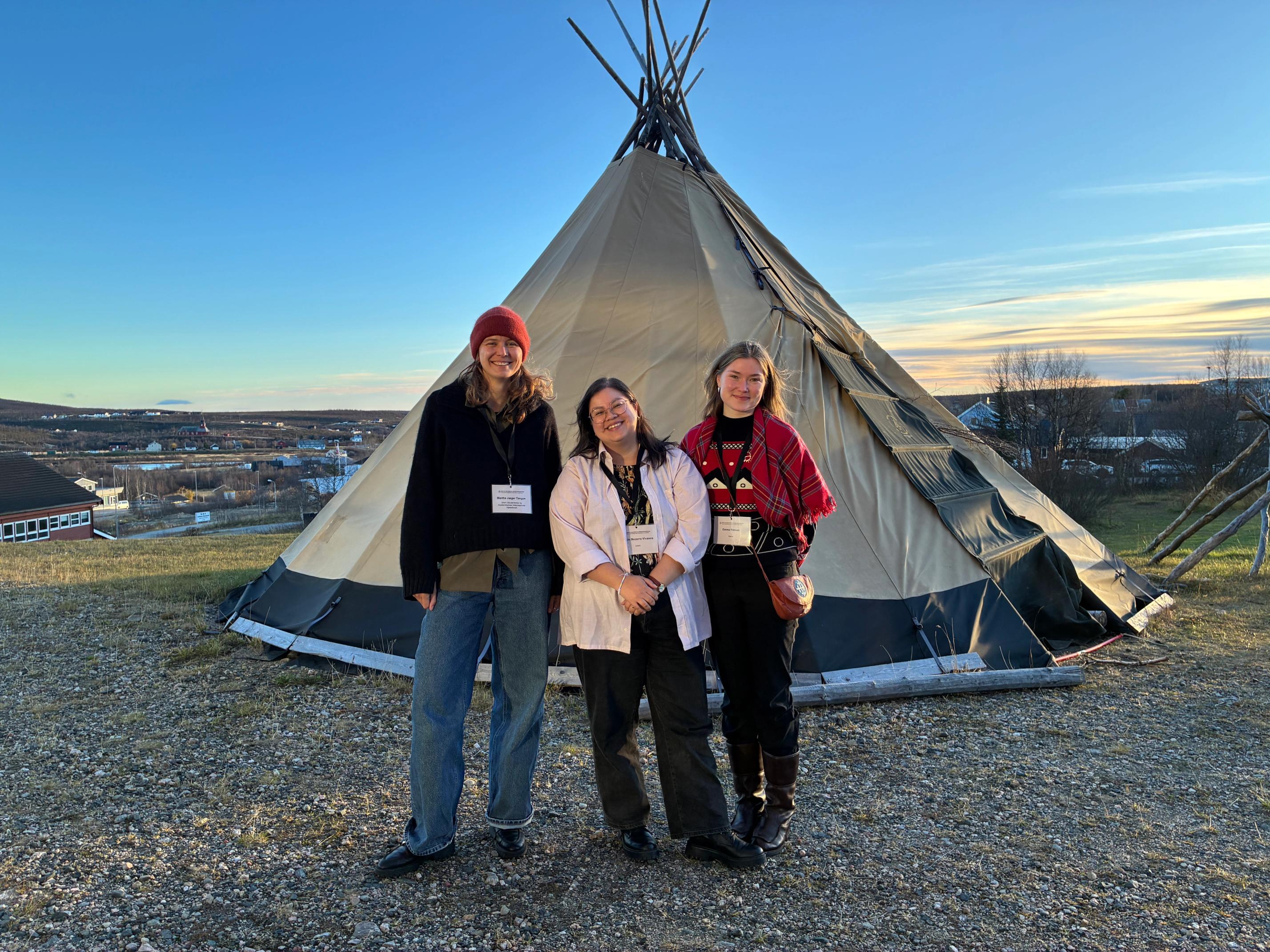 Three smiling people stand in front of a large conical tent, with a town visible in the background under a golden sky.