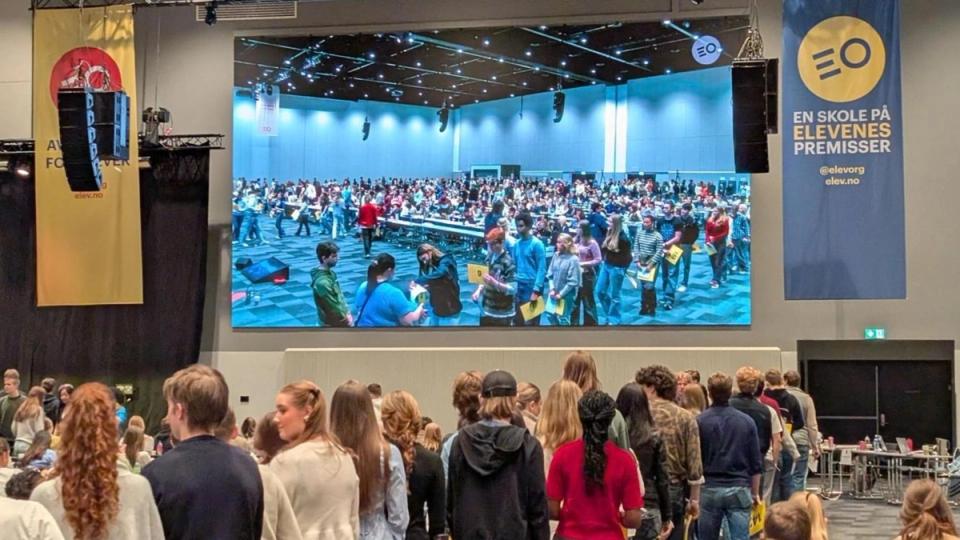 A large screen at a student event shows a room full of people, as other students line up in the foreground.