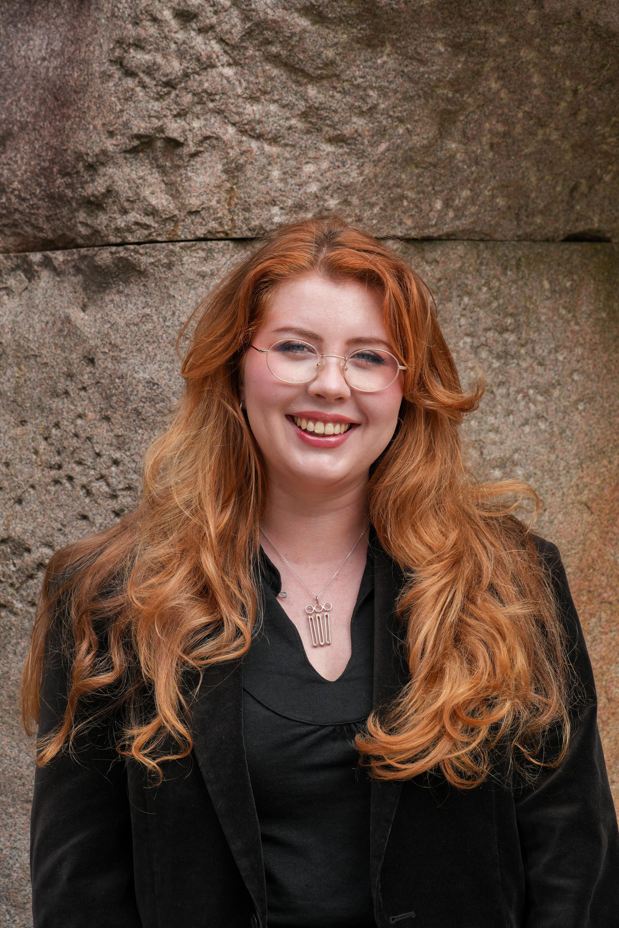 Smiling young woman with long, wavy red hair, glasses, a black blazer, and a silver pendant, against a stone wall.