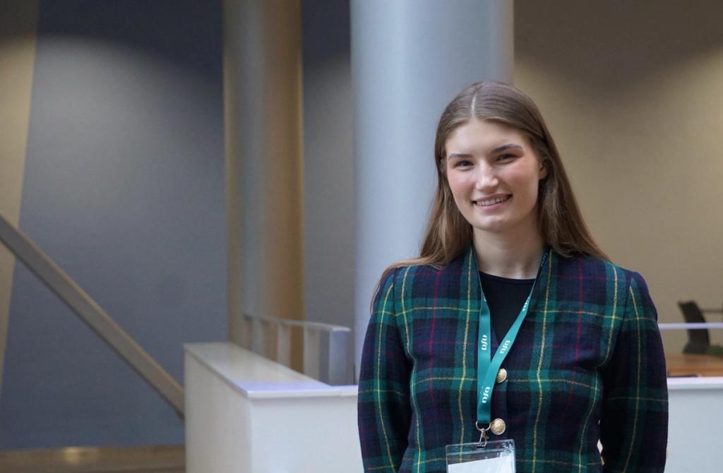 A smiling young woman with long brown hair wears a green and blue plaid jacket and a teal lanyard with an ID badge, standing in an indoor space.