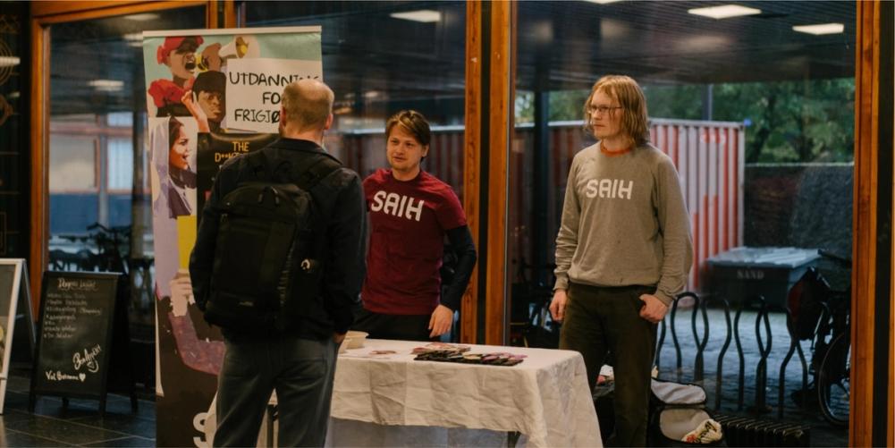 Two men in "SAIH" shirts stand behind a table, talking to a third man with his back to the camera.