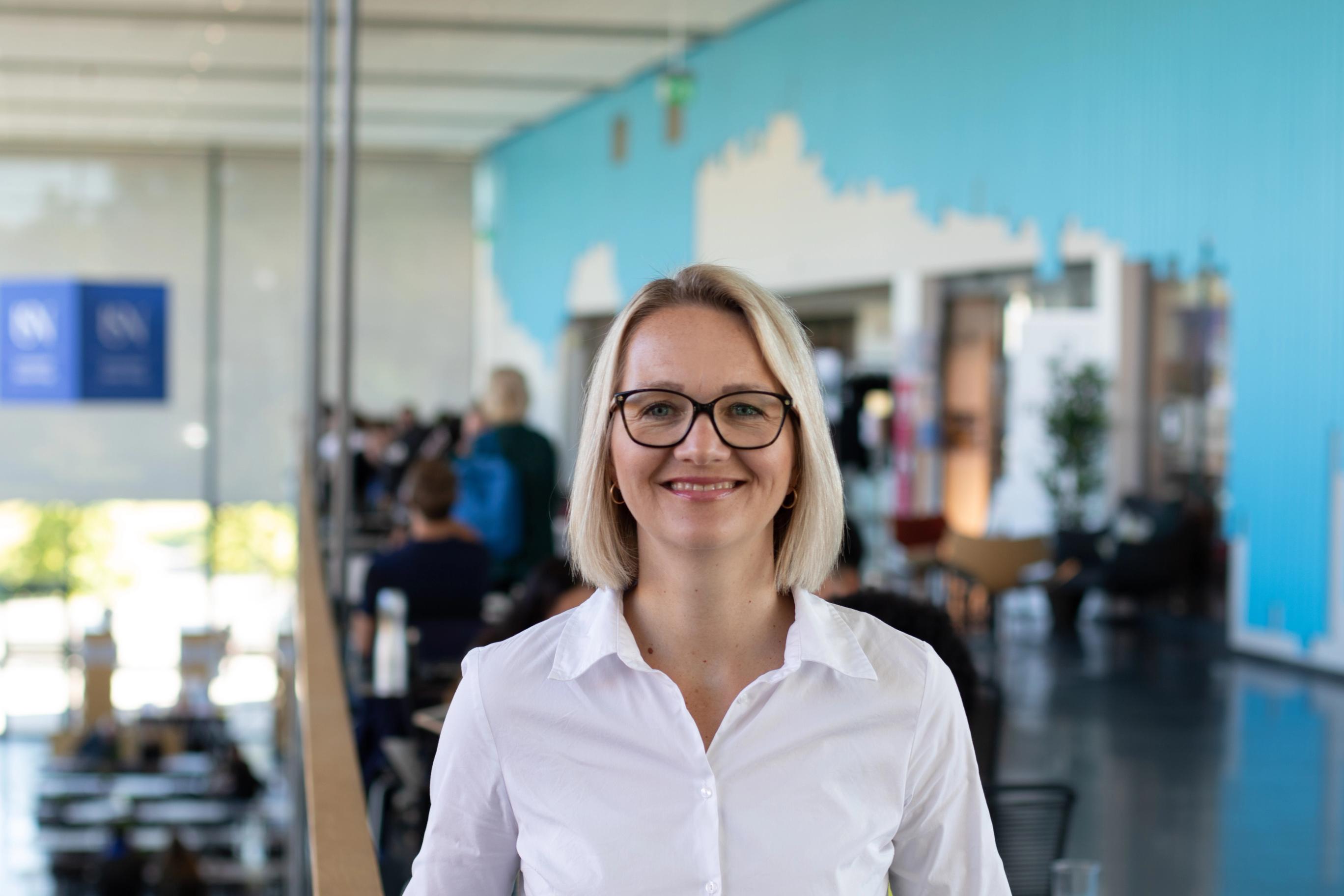 Smiling blonde woman in glasses and a white shirt in a modern office.