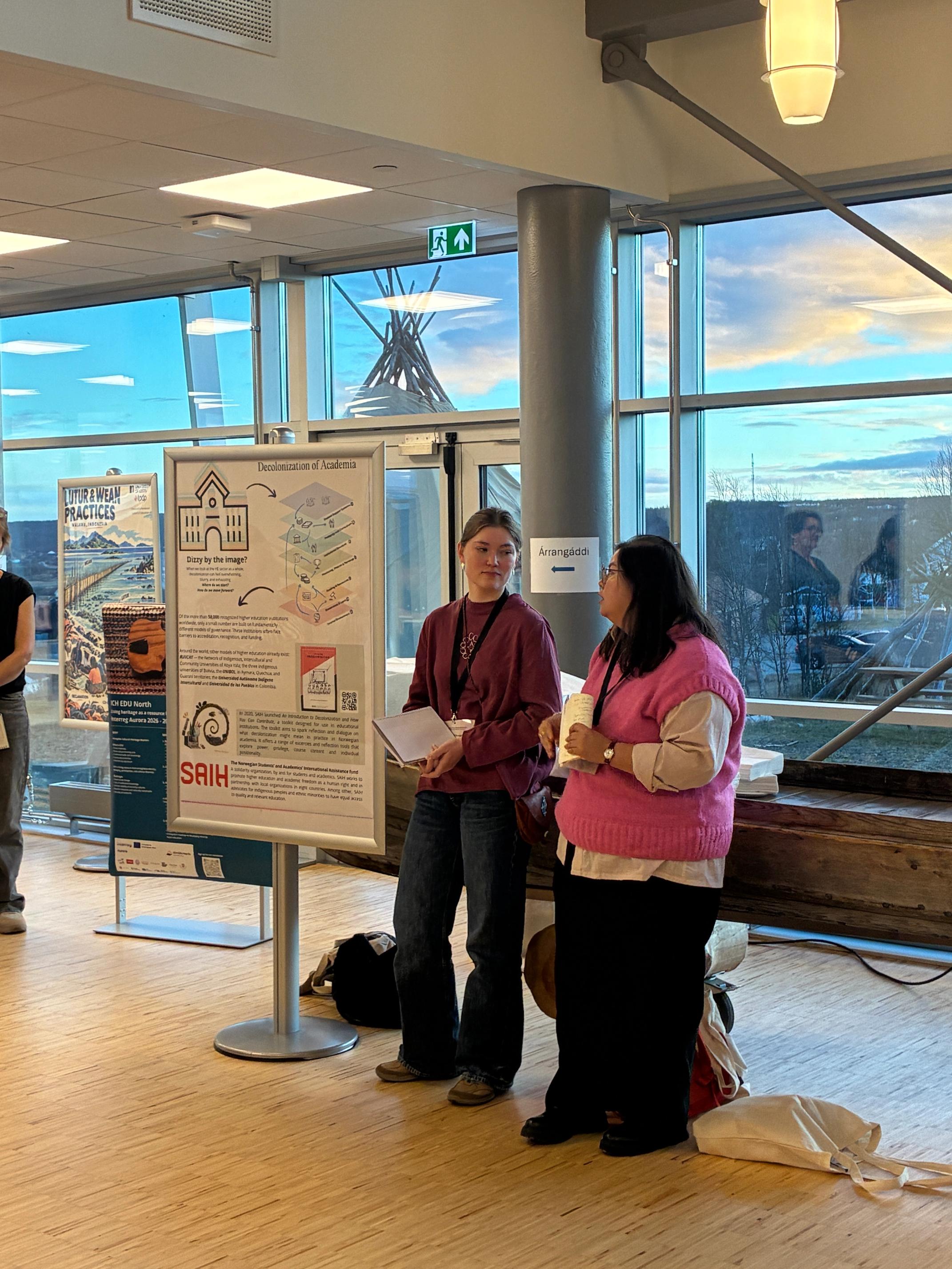 Two women stand next to a poster titled "Decolonization of Academia" in a modern building with large windows showing a teepee-like structure outside.