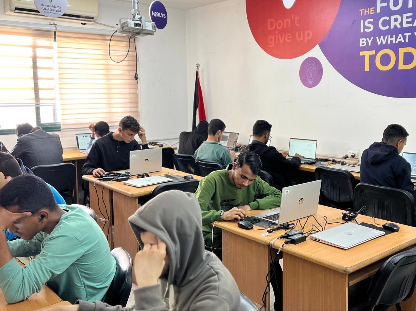 A classroom of young men working on individual laptops, with motivational wall art and a Palestinian flag visible in the background.