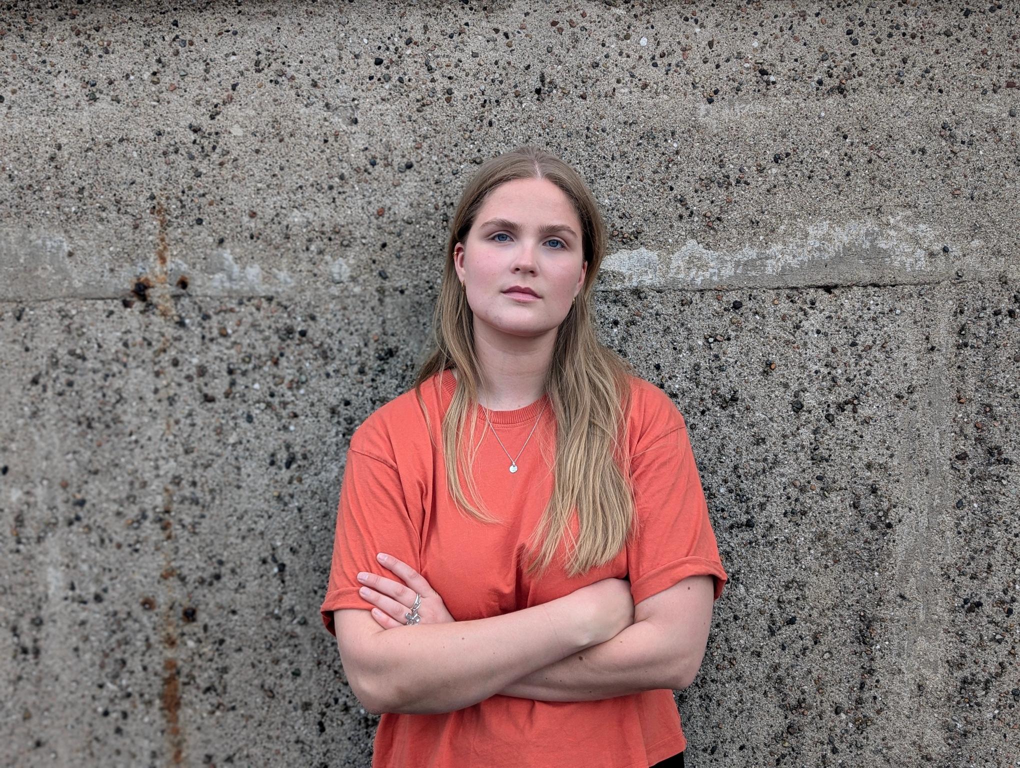 A young woman with blonde hair, an orange t-shirt, and crossed arms stands against a concrete wall, looking forward.