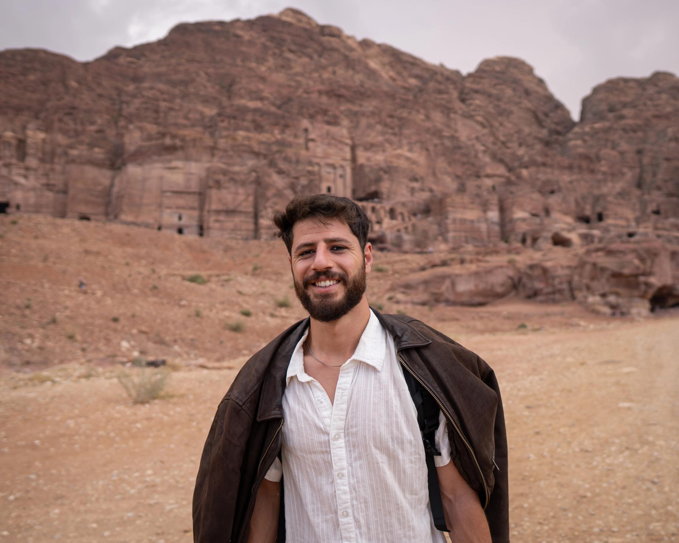 Smiling man in front of ancient rock-cut structures in Petra's red desert mountains.
