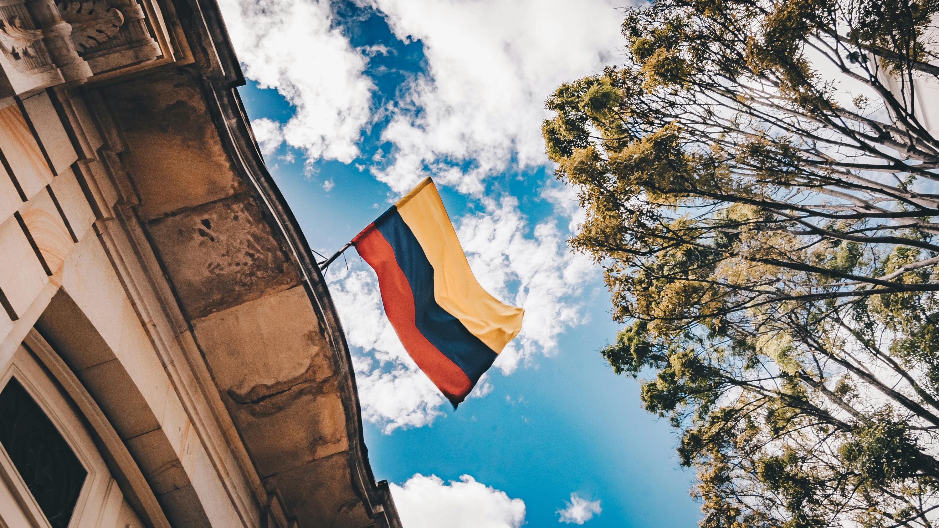 Colombian flag flying from a stone building with trees and a cloudy blue sky.