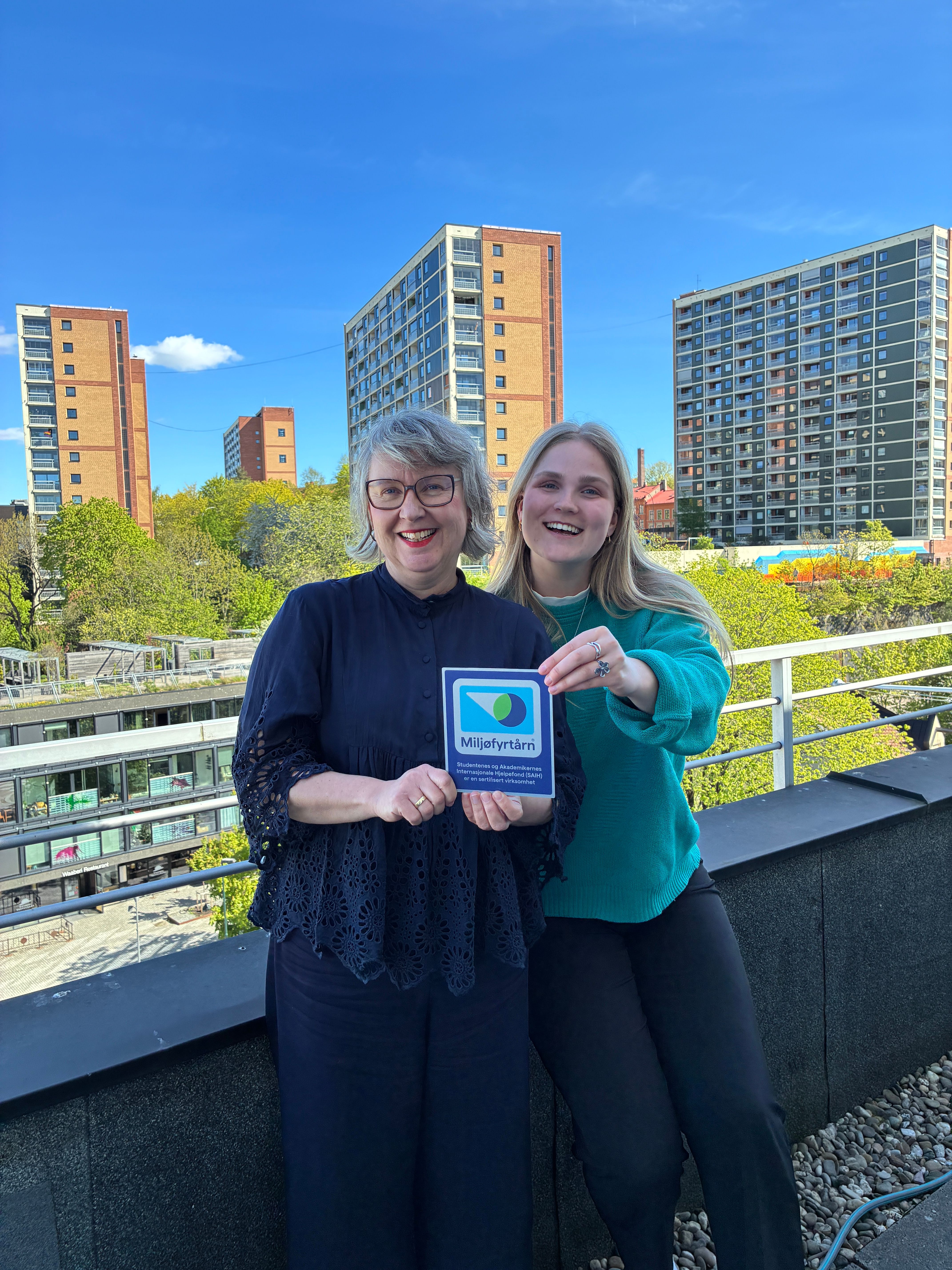 Two smiling women hold a "Miljöfyrtårn" sign on a rooftop with city buildings and trees in the background.