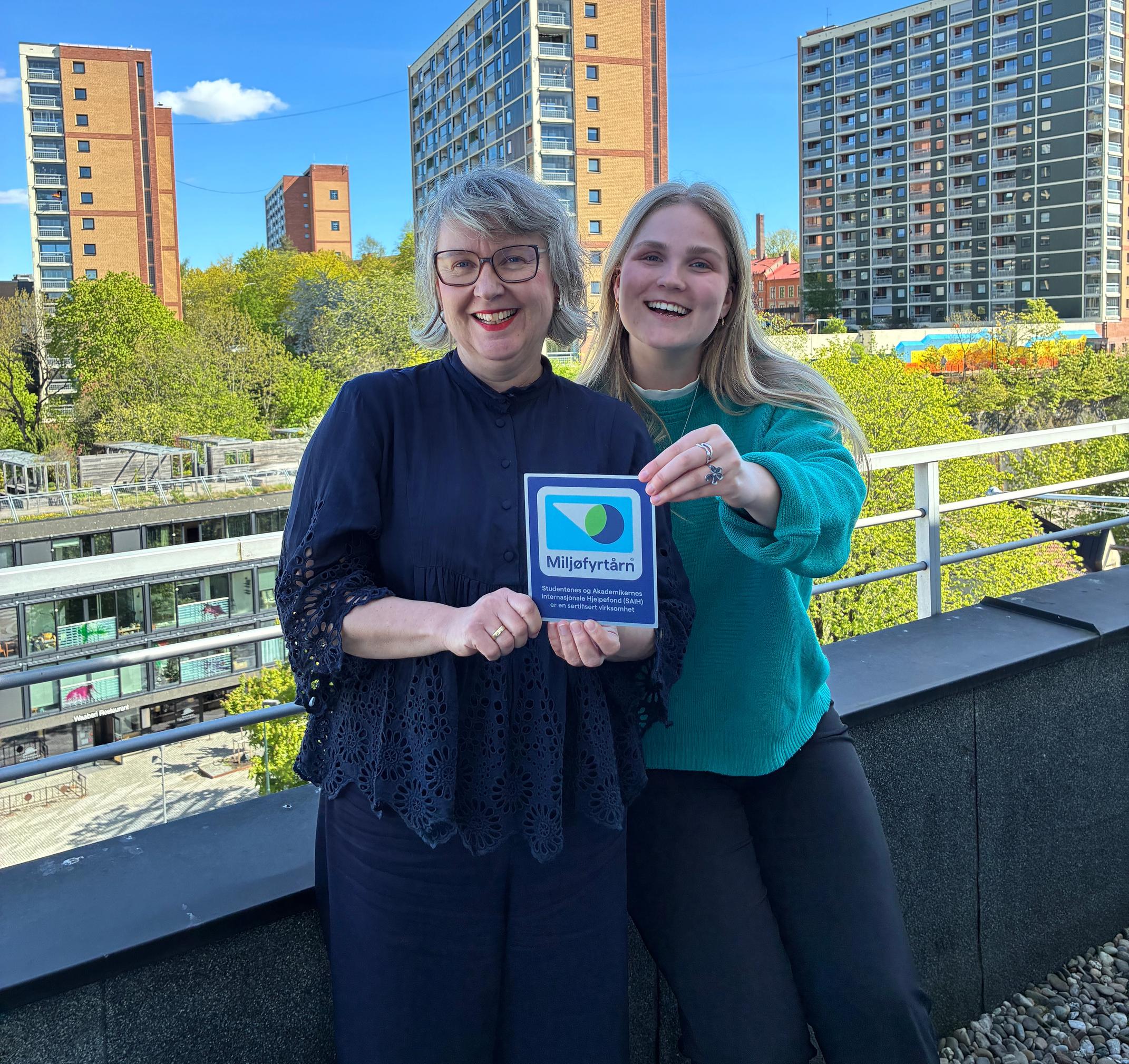Two smiling women hold a "Miljöfyrtårn" sign on a rooftop with city buildings and trees in the background.