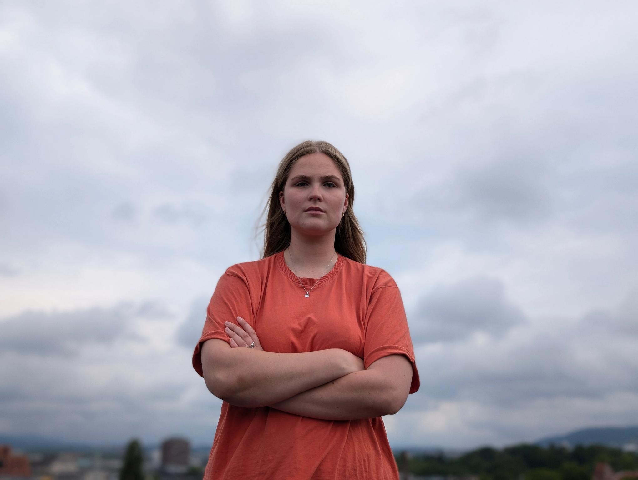 A woman in an orange t-shirt with crossed arms and a serious expression stands against a cloudy sky.