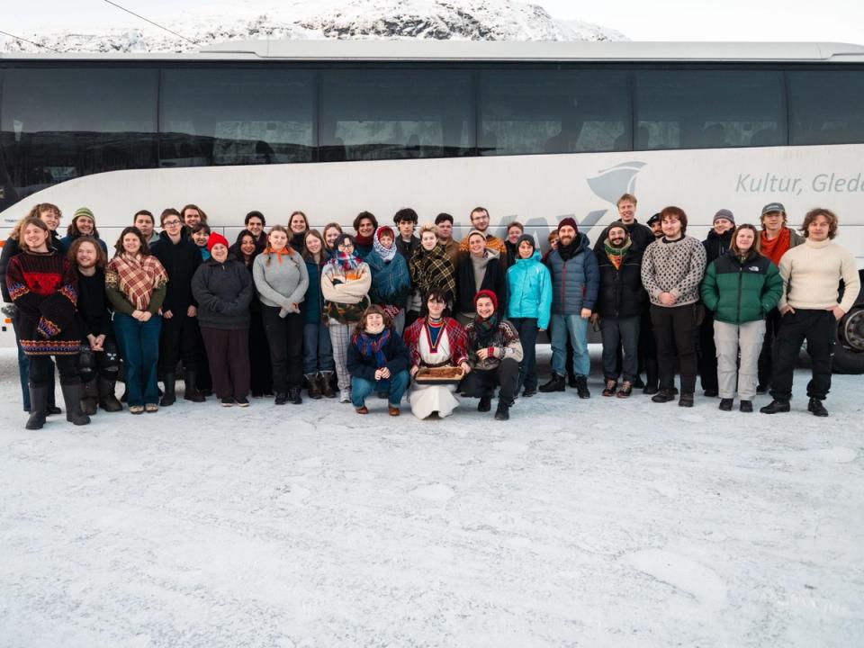 A large group of people stand on snowy ground in front of a white bus with mountains in the background.