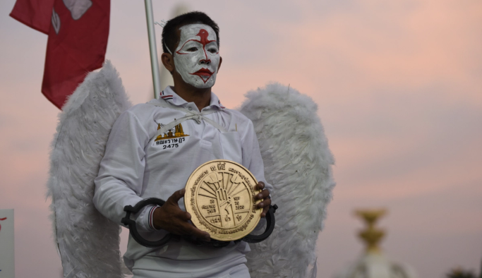 A person wearing white angel wings, a white and red painted face, and shackles, holds a large golden disc.