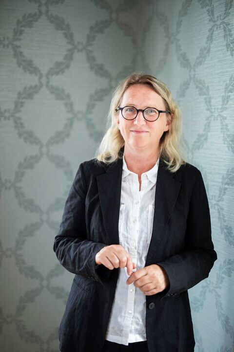 A blonde woman in glasses, a black blazer, and a white shirt smiles with hands clasped against a patterned wall.