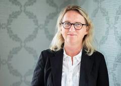 A blonde woman in glasses, a black blazer, and a white shirt smiles with hands clasped against a patterned wall.