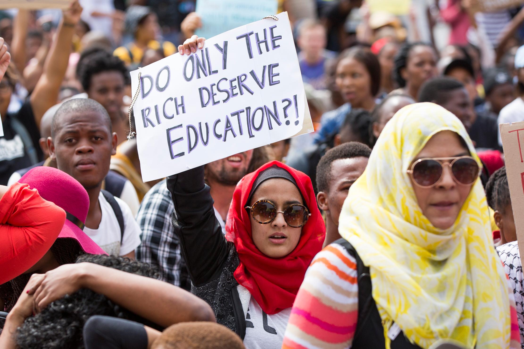 En kvinne stående i en stor folkemengde holder et skilt som stiller spørsmål om hvorvidt det kun er de rike som fortjener utdanning. 