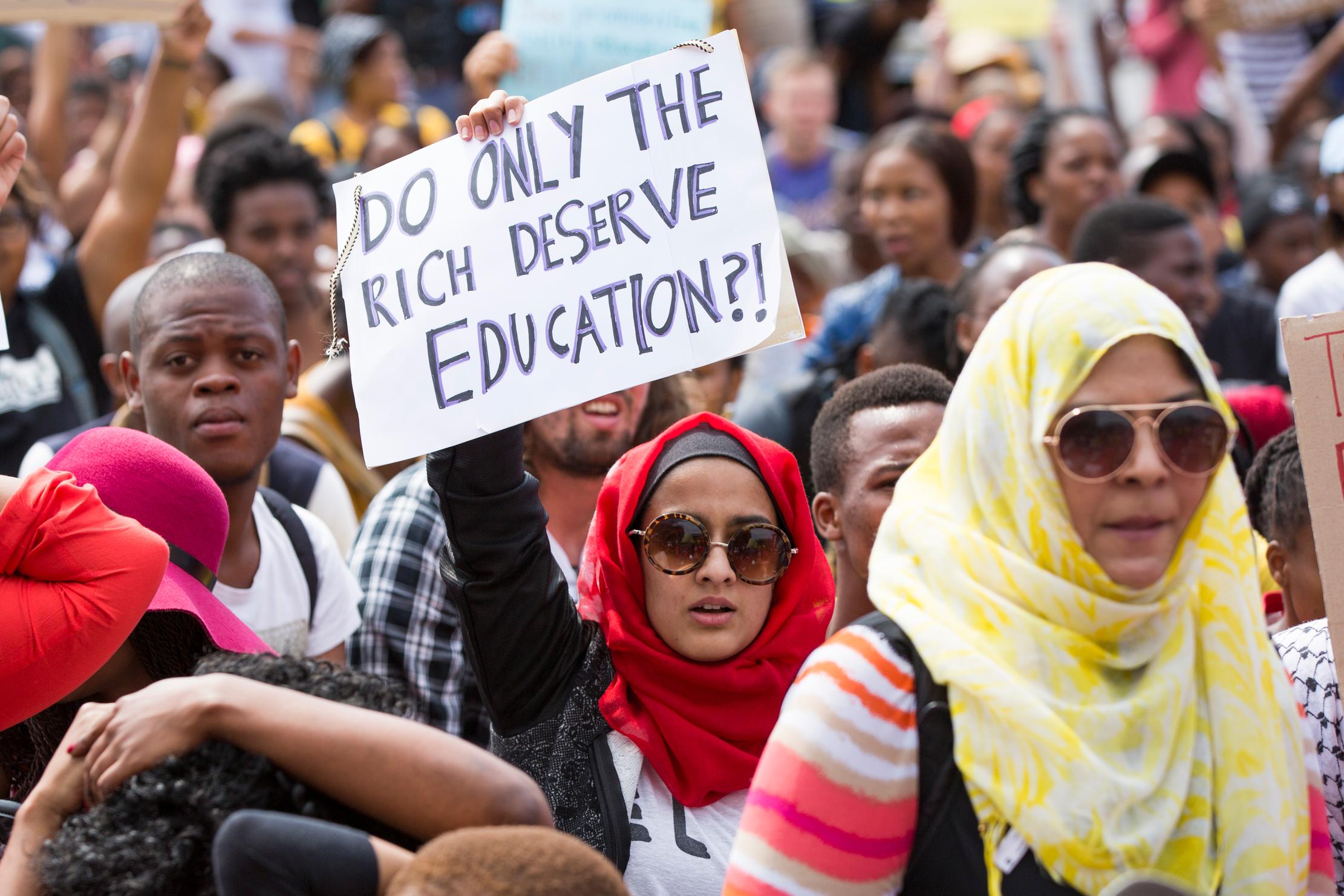 A group of people demonstrating, one with a sign questioning whether only the rich should have access to education.